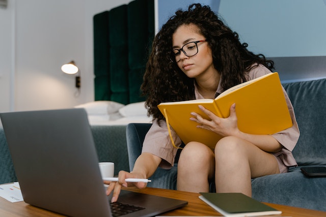 Person with long hair and glasses sitting on a couch, holding a notebook and using their laptop